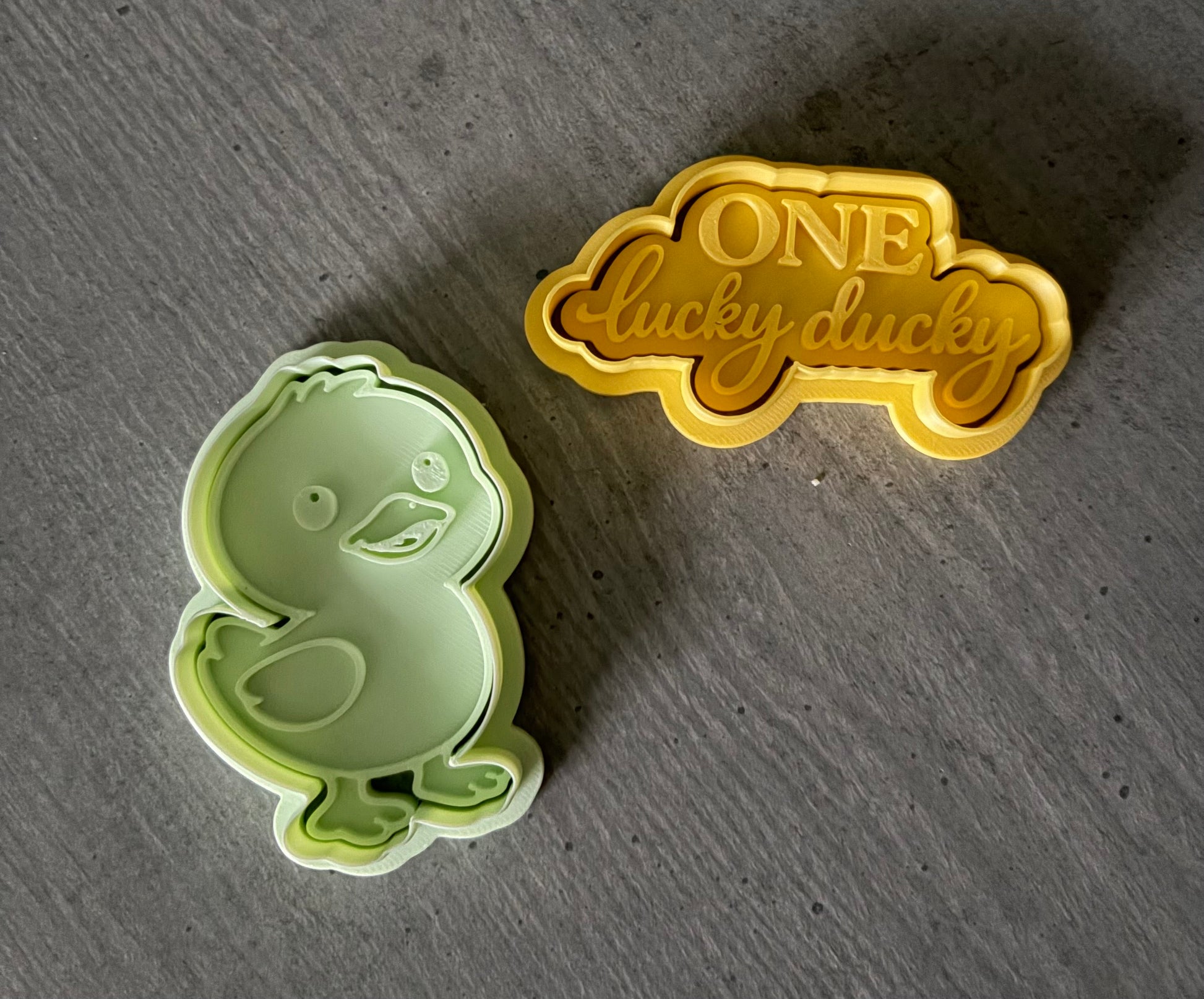 Green duck-shaped cookie cutter and gold 'ONE lucky duck' stamp on a gray surface
