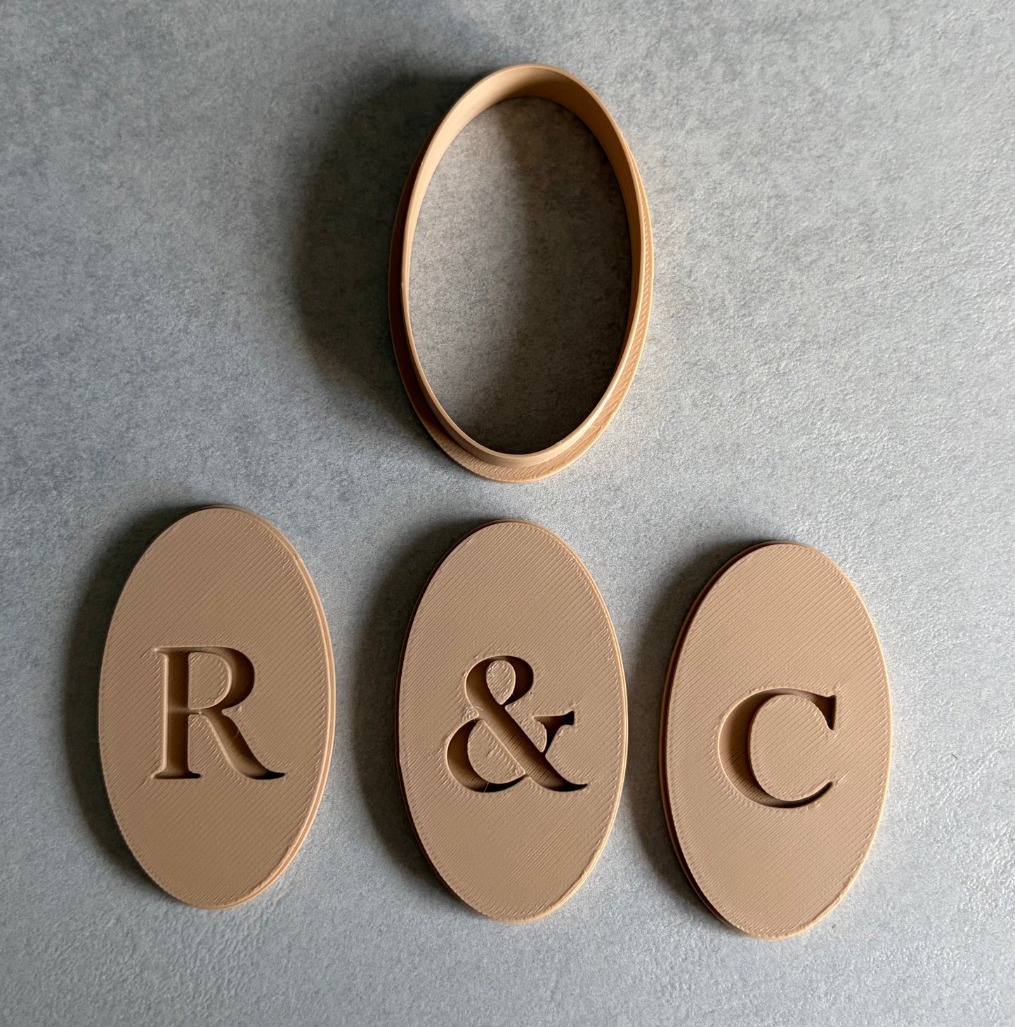 Three oval beige monogram pieces with engraved letters R, &, and C, plus a separate oval ring piece, arranged on a gray textured background.