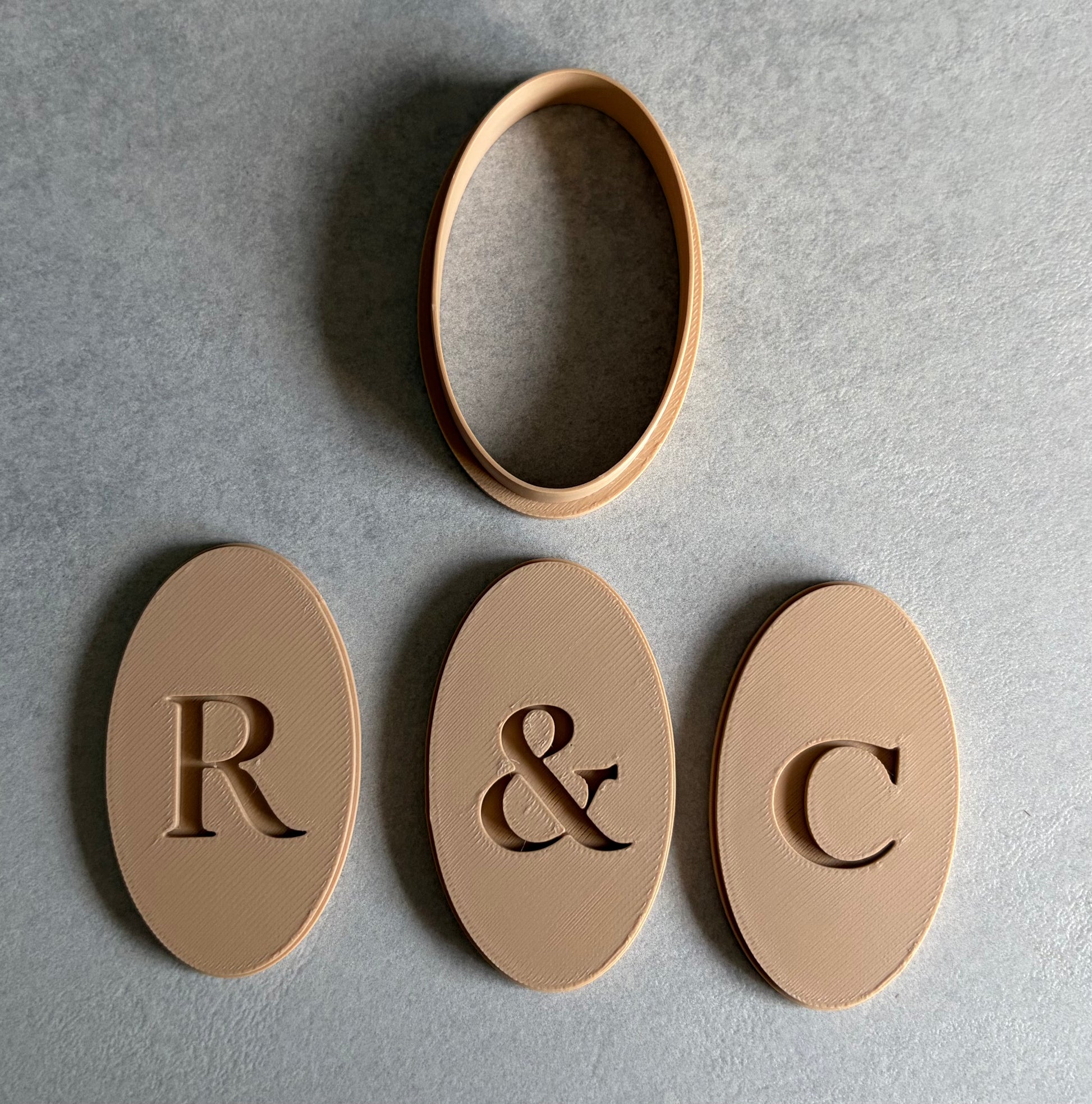 Three oval beige monogram pieces with engraved letters R, &, and C, plus a separate oval ring piece, arranged on a gray textured background.