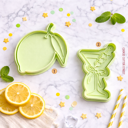 Two green cookie cutters shaped like lemons and a cocktail glass on a marble surface with lemon slices and mint leaves.