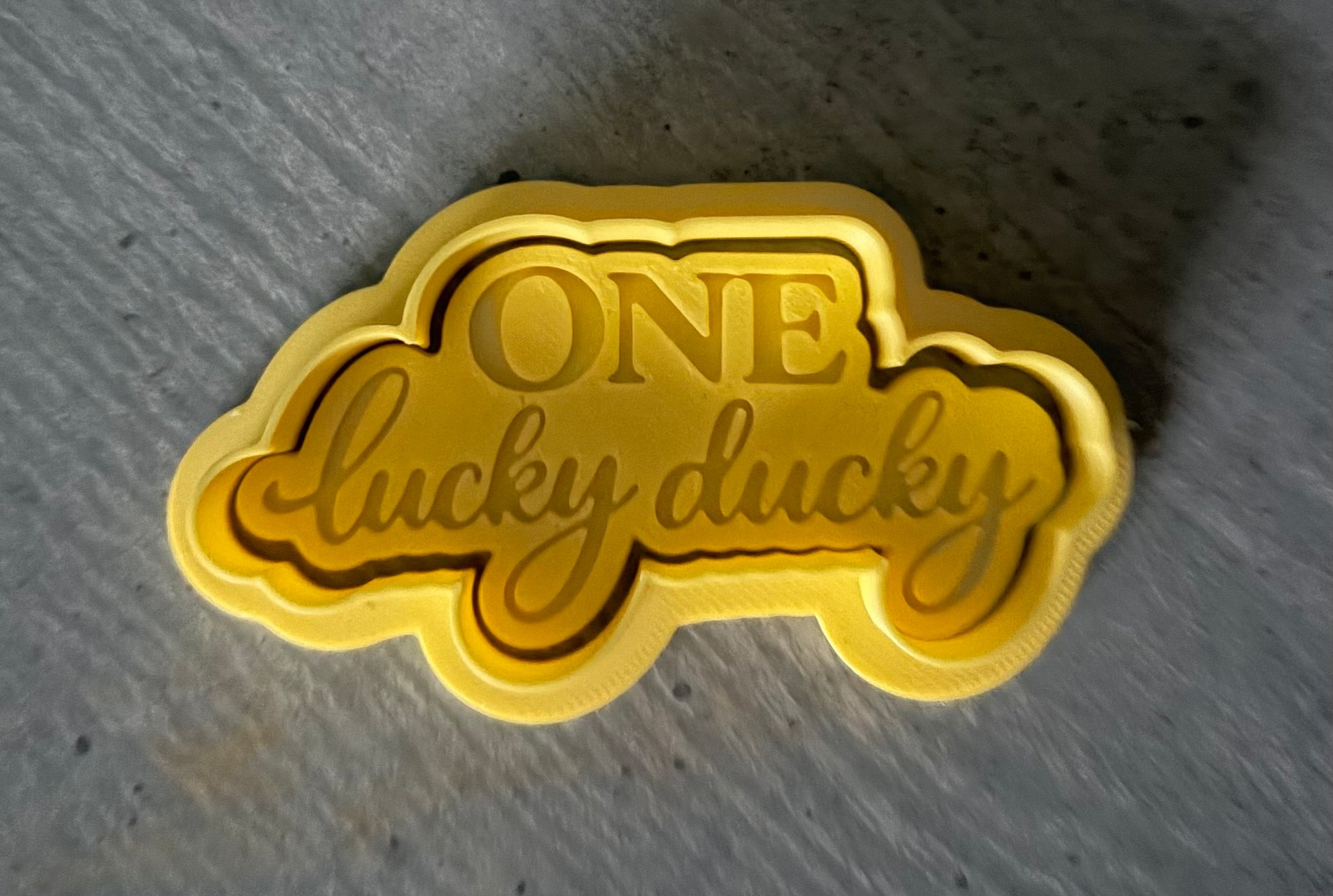 Gold 'ONE lucky duck' cookie cutter on a textured gray background