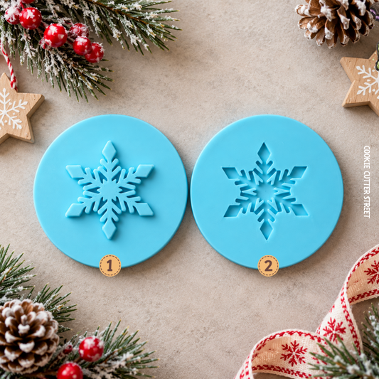 Two round blue cookie cutters with snowflake designs, labeled 1 and 2, displayed on a neutral beige surface with festive Christmas decorations including pine branches, red berries, and wooden star ornaments.