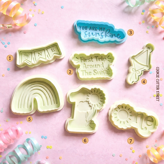 Baby-themed cookie cutter and imprint stamp set featuring “One” designs including rainbow, sunshine and birthday text for first birthday cookies.