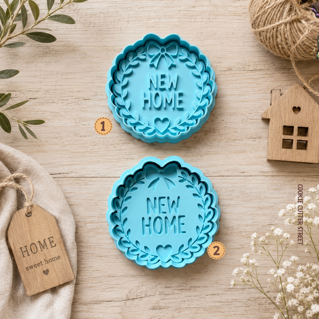 Two blue scalloped cookie cutters with 'NEW HOME' embossed in the center, arranged on a light wood surface with decorative props including twine, flowers, a wooden house cutout, and a fabric tag.